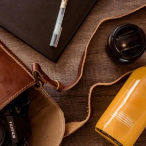 Stylish flat lay of leather bag, camera lens, and refreshing orange beverage on wooden surface.